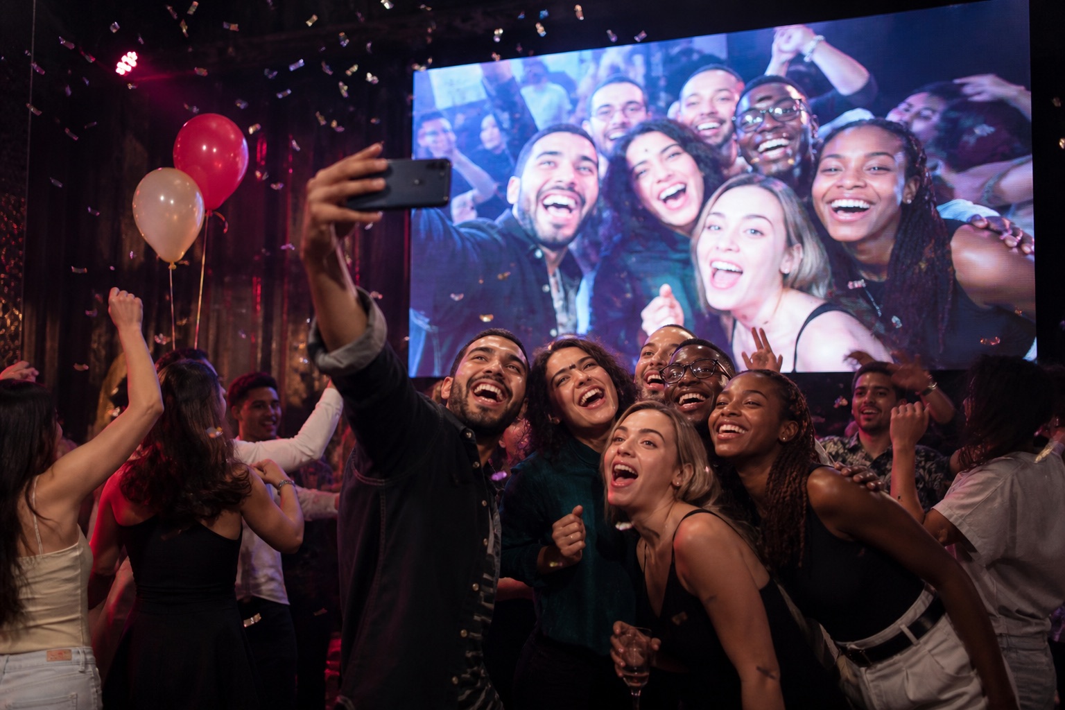 Friends taking a playful selfie at a neon party while it appears on a large LED screen